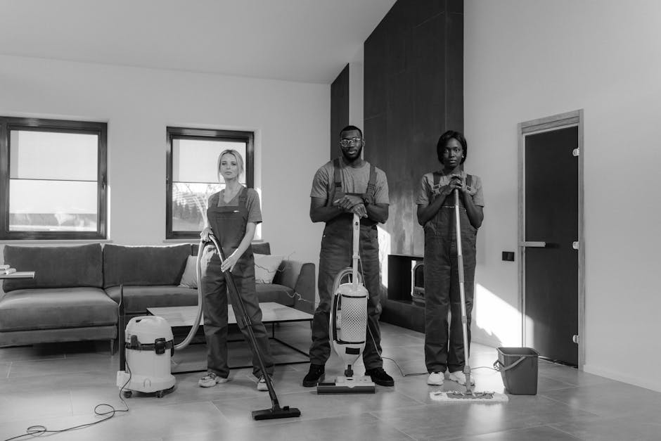 Three professional cleaners from Cleaners W5 stand in a spacious, well-lit living room preparing for surface cleaning. The room features a large L-shaped fabric sofa with a low wooden coffee table nearby, and a modern entertainment console with a small microwave on it. The wooden floor is smooth and clean, reflecting light from two large windows with partially closed blinds, revealing an outdoor view. Each cleaner is dressed in dark uniforms with aprons; the female to the left holds a vacuum hose, the male in the middle is ready with a canister vacuum, and the female on the right holds a mop and a bucket, indicating ongoing deep cleaning and sanitisation activities. The walls are painted white, complemented by a dark accent wall behind the cleaners. The scene emphasizes thorough domestic cleaning, surface hygiene, and attention to detail, showcasing Cleaners W5’s commitment to quality cleaning services for a spotless and healthy living environment.