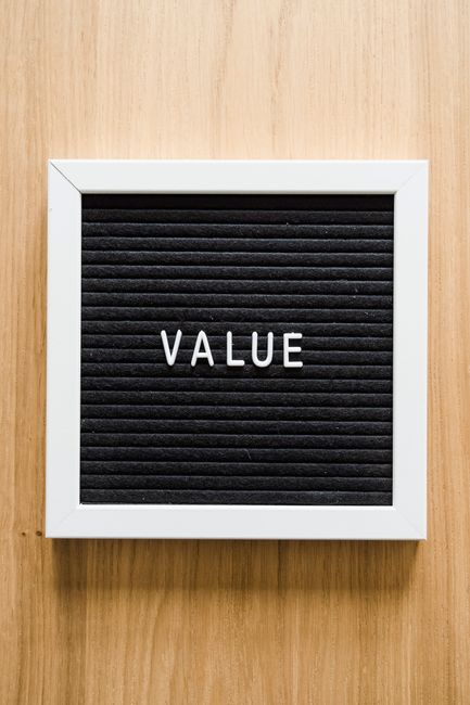 A close-up view of a square black felt letterboard with white plastic frame resting on a light wooden surface. The word 'VALUE' is spelled out in white plastic letters centered on the board. The surface of the wooden background appears smooth and clean, illuminated by soft, natural lighting that highlights the texture of the wood and the contrast between the dark board and white frame. This image emphasizes clarity and cleanliness, relevant to the importance of transparent and honest service quotes in the context of domestic cleaning, as discussed on the page 'Avoid hidden fees in Ealing cleaning quotes what to check, W5' by Cleaners W5.