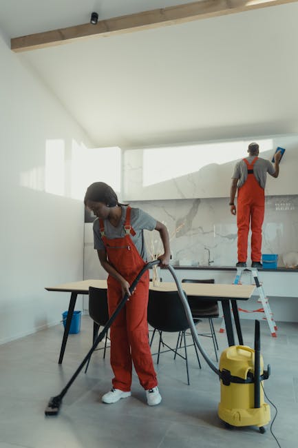 Three professional cleaners from Cleaners W5 stand in a spacious, well-lit living room preparing for surface cleaning. The room features a large L-shaped fabric sofa with a low wooden coffee table nearby, and a modern entertainment console with a small microwave on it. The wooden floor is smooth and clean, reflecting light from two large windows with partially closed blinds, revealing an outdoor view. Each cleaner is dressed in dark uniforms with aprons; the female to the left holds a vacuum hose, the male in the middle is ready with a canister vacuum, and the female on the right holds a mop and a bucket, indicating ongoing deep cleaning and sanitisation activities. The walls are painted white, complemented by a dark accent wall behind the cleaners. The scene emphasizes thorough domestic cleaning, surface hygiene, and attention to detail, showcasing Cleaners W5’s commitment to quality cleaning services for a spotless and healthy living environment.