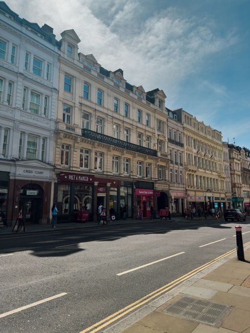 Image of a street in Ealing Broadway showing a row of multi-storey commercial buildings with classic architectural details, including large windows and ornate facades, under a partly cloudy sky. The ground floor features various retail shops and cafes with signage, while pedestrians walk along the sidewalk. The street has parked cars and the road surface appears clean and well-maintained, with visible road markings and bollards for pedestrian safety. Natural daylight illuminates the scene, highlighting the tidy appearance of the area. This setting demonstrates the local urban environment where professional cleaning services by Cleaners W5 could be utilized for maintaining the hygiene and appearance of commercial premises.