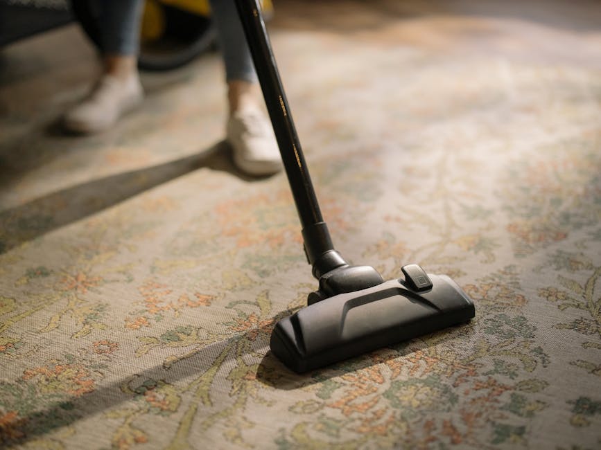 Close-up of a vacuum cleaner head actively cleaning a traditional patterned area rug with floral motifs in a well-lit room. The vacuum is black with a slightly textured surface, and its head is flat and wide, covering a surface area of the carpet. Part of the vacuum handle is visible, held by a person wearing white footwear and dark trousers, suggesting a professional cleaning process. The rug appears clean and free of dust or debris, with a subtle sheen indicating recent or ongoing deep cleaning. The ambient lighting highlights the vibrant colours and intricate details of the rug's design, and the overall scene reflects a professional approach to surface cleaning and upholstery maintenance, consistent with the services offered by Cleaners W5 in the Ealing Broadway area.