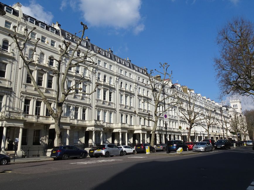 Image of a street in Ealing Broadway showing a row of multi-storey commercial buildings with classic architectural details, including large windows and ornate facades, under a partly cloudy sky. The ground floor features various retail shops and cafes with signage, while pedestrians walk along the sidewalk. The street has parked cars and the road surface appears clean and well-maintained, with visible road markings and bollards for pedestrian safety. Natural daylight illuminates the scene, highlighting the tidy appearance of the area. This setting demonstrates the local urban environment where professional cleaning services by Cleaners W5 could be utilized for maintaining the hygiene and appearance of commercial premises.