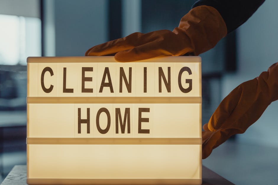 A person wearing orange cleaning gloves is adjusting a lit-up sign with black letters on a white background that reads 'CLEANING HOME.' The sign is placed on a surface in a well-lit interior space, possibly a kitchen or living area, with tiled surfaces and visible windows in the background. The display emphasizes the importance of surface cleaning and sanitisation in domestic environments, aligning with the Northfields end of tenancy cleaning guide for Ealing landlords, W5, provided by Cleaners W5.