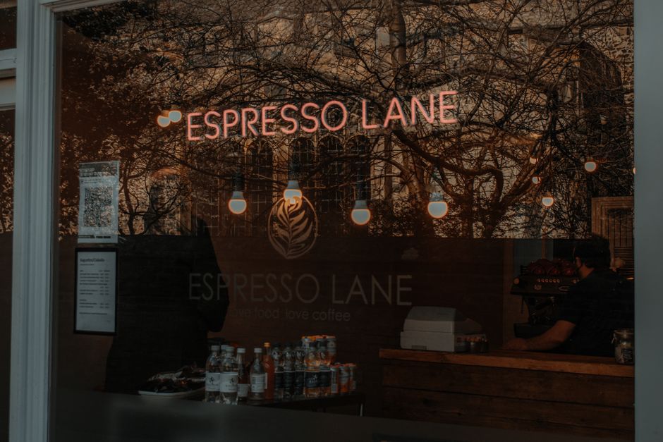 A view through the window of Espresso Lane, a coffee shop on Pitshanger Lane in Ealing, W5. The shop's illuminated pink sign reading 'ESPRESSO LANE' is visible, with hanging pendant lights inside creating a warm ambient glow. The interior features a wooden counter with visible coffee equipment, and various bottled beverages are arranged on a tray near the window. Reflections of trees and the surrounding street can be seen on the glass, which appears clean and clear. The scene suggests a well-maintained commercial space with a welcoming atmosphere, possibly recently cleaned or deep cleaned by professionals such as those from Cleaners W5.