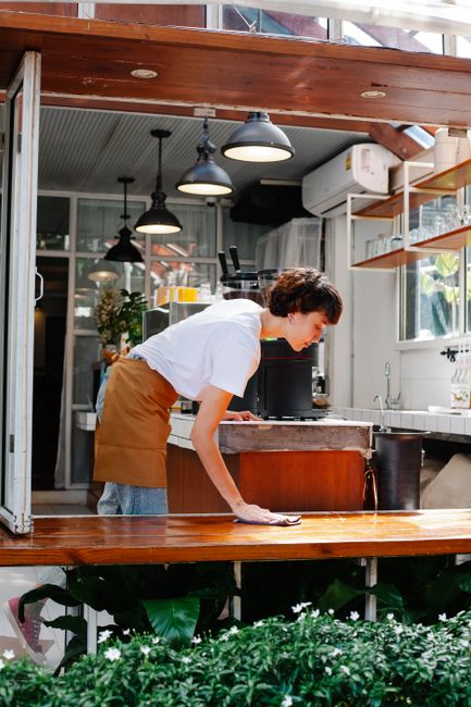 A young woman dressed in a black long-sleeve shirt, a white apron, and patterned tights is standing at the entrance of a shop or café on Pitshanger Lane. She holds a cleaning cloth and a spray bottle, indicating ongoing surface cleaning or sanitisation. The interior features shelves with various containers and a glass display, visible behind her, reflecting natural light. The doorway has a glass panel and a partially open door, with a tiled exterior wall nearby. The scene emphasizes cleanliness and maintenance typical of commercial establishments in Ealing, W5, as stained or dusty surfaces are absent, and the area appears tidy and well-kept, consistent with professional cleaning services by Cleaners W5.
