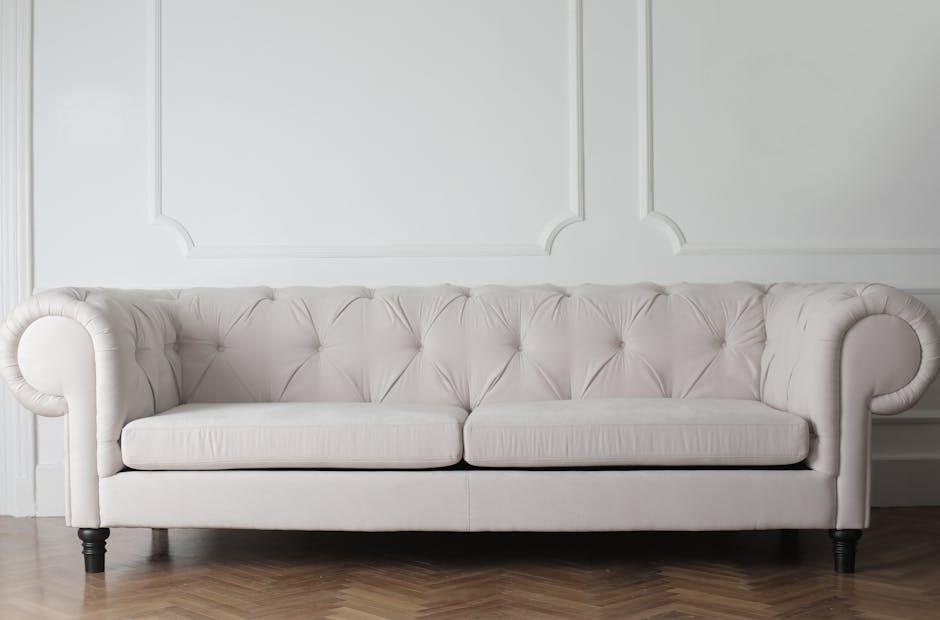 A pristine, white upholstered sofa with tufted backrest and rolled armrests sits in a well-lit living room with white paneled walls. The wooden floor beneath is visible, adding a warm tone to the scene. The sofa appears thoroughly cleaned, with no visible dust or stains, reflecting professional surface cleaning and maintenance by Cleaners W5, specializing in deep cleaning and sanitisation for residential spaces, including upholstery cleaning and stain removal in Ealing, W5.