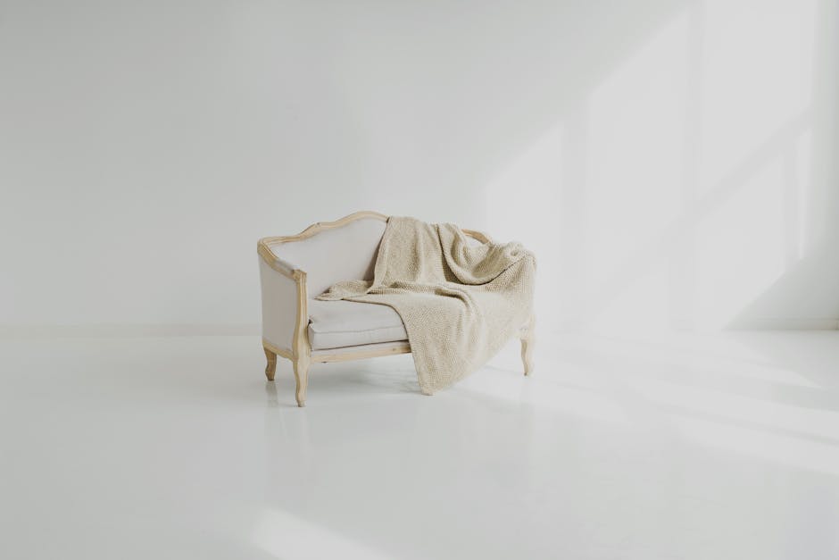 A vintage-style white armchair with wooden legs and ornate detailing sits in the corner of a bright, minimalist room with white walls and a smooth white floor. A beige knitted throw blanket is casually draped over the armrest and seat of the chair. The room is filled with natural light coming from a large window on the right, casting soft shadows and illuminating the clean, uncluttered space. The setting emphasizes a sense of cleanliness and simplicity, which aligns with professional domestic cleaning and surface sanitisation practices highlighted by Cleaners W5 in their Pitshanger Park upholstery cleaning and stain removal services in Ealing, W5.